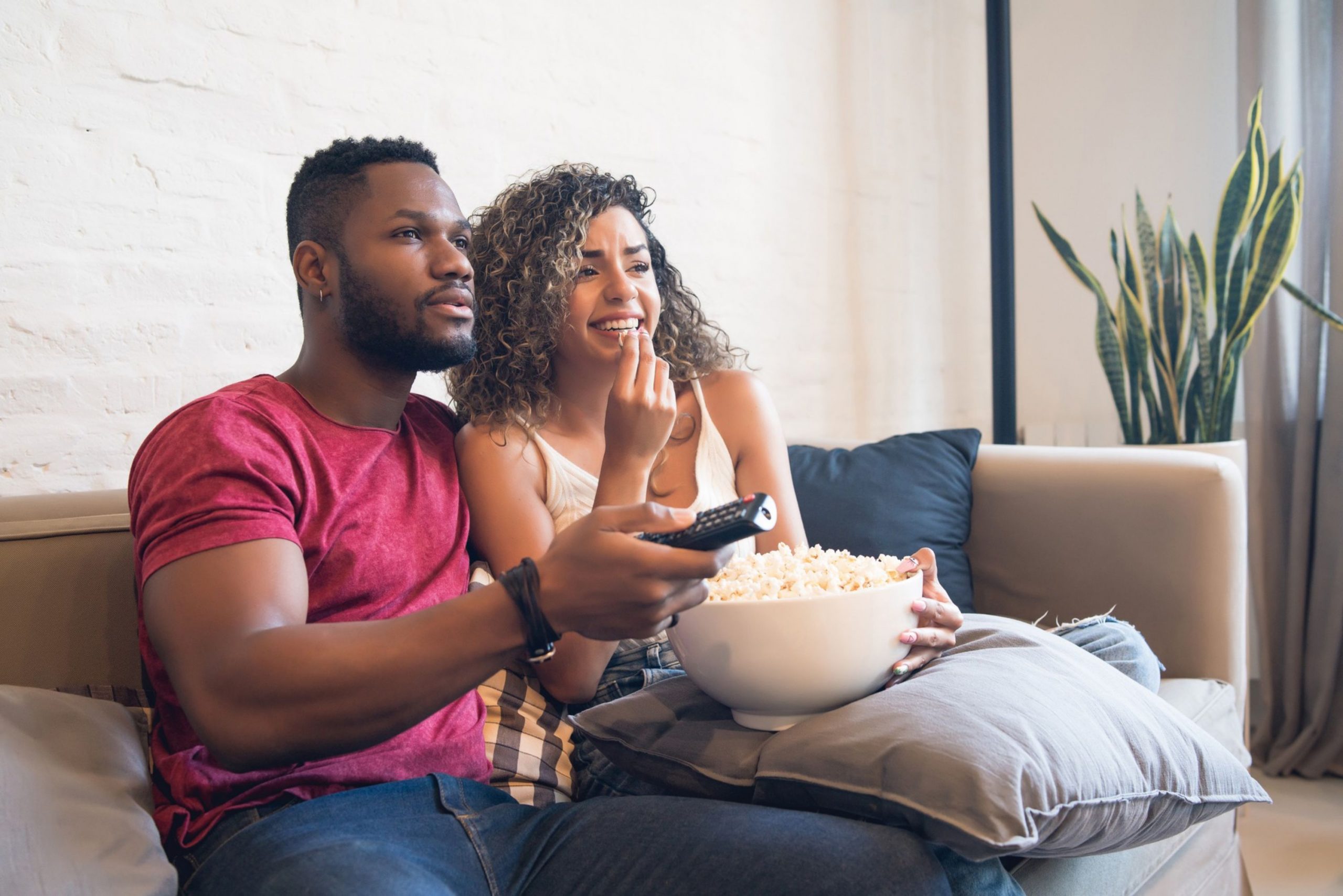 Couple sitting on the sofa watching TV whilst eating popcorn.